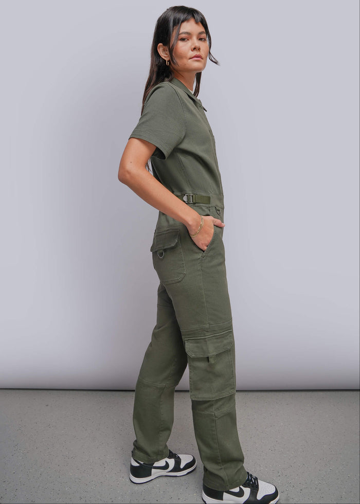 S=A woman modeling an olive green short-sleeve utility jumpsuit with cargo pockets and an adjustable waist, paired with black and white sneakers, standing against a light gray studio background.