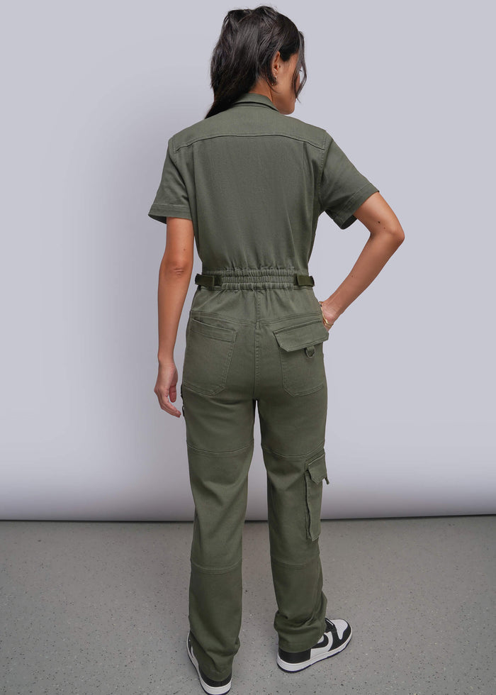 S=A woman shown from the back wearing an olive green short-sleeve utility jumpsuit with cargo pockets and an adjustable waist, paired with black and white sneakers, against a light gray studio background.