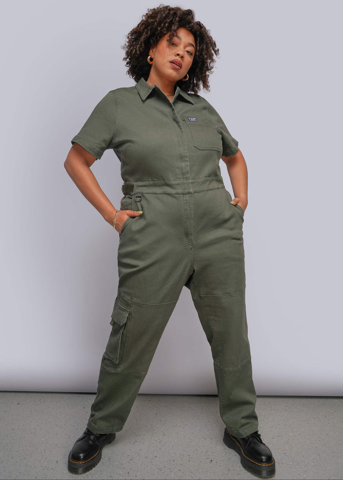 1X=A woman modeling an olive green short-sleeve utility jumpsuit with cargo pockets, standing against a light gray studio background.
