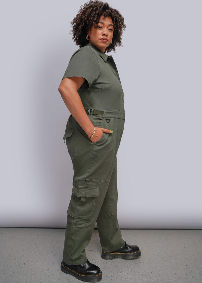 1X=A woman modeling an olive green short-sleeve utility jumpsuit with cargo pockets and a belt, paired with black platform shoes, against a light gray studio background.