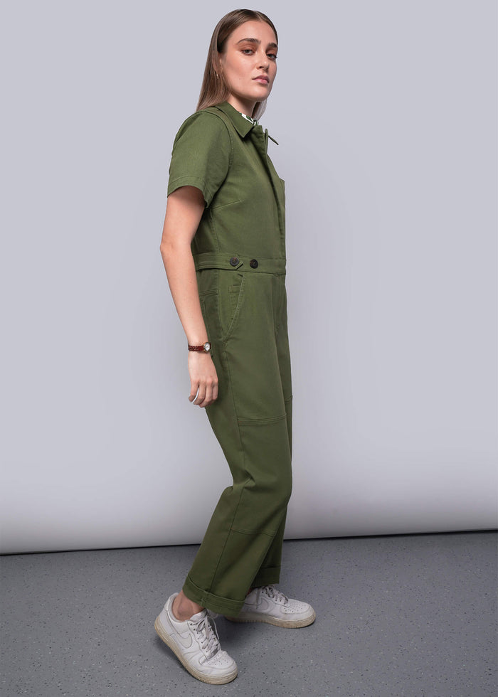 S=Woman wearing a green jumpsuit standing against a plain background