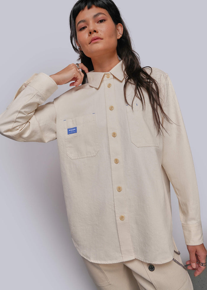 S=A model wearing a light beige long-sleeve button-up shirt with a small blue brand patch on the chest pocket, paired with matching beige trousers, against a light gray studio background.