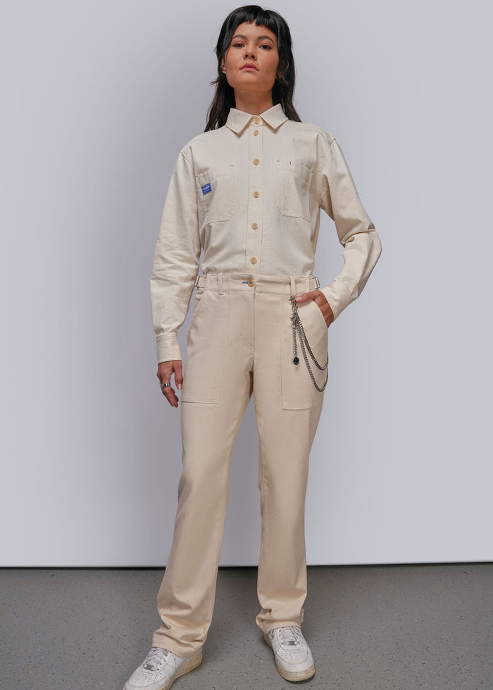 S=A model stands against a light gray studio wall wearing a beige utility-style shirt and matching trousers with a chain detail at the waist, paired with white sneakers.
