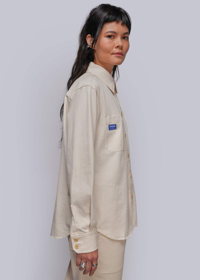 S=A model wearing a light beige linen button-up shirt with a small blue brand patch on the chest pocket, paired with matching beige trousers, set against a plain light gray studio background.