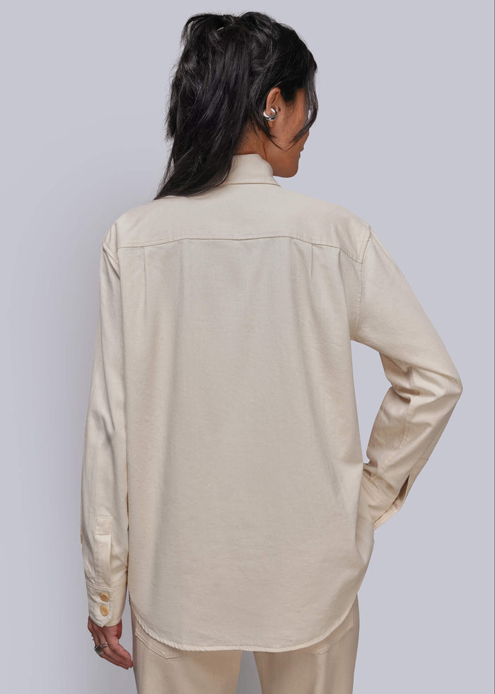 S=A woman shown from the back wearing a light beige long-sleeve button-up shirt and matching beige pants against a light gray studio background.
