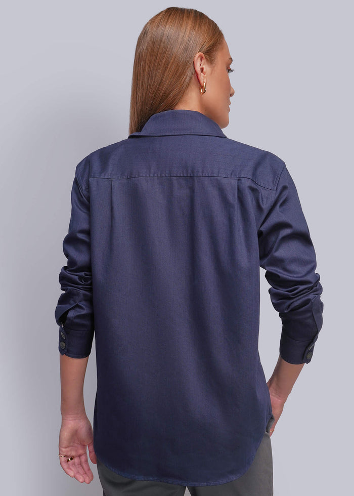 S=A woman shown from the back wearing a dark navy blue long-sleeve button-up shirt and gray trousers, standing against a solid light gray studio background.