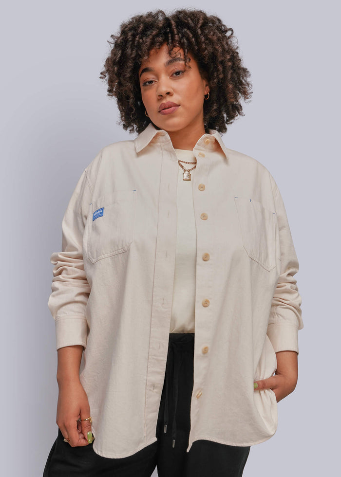 1X=A woman with curly hair wearing a light beige button-up shirt with a small blue logo patch on the chest pocket, paired with black pants, against a solid light gray background.
