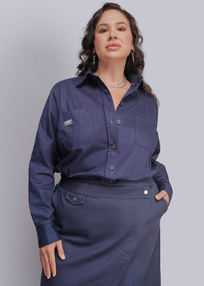 1X=A woman modeling a navy blue long-sleeve button-up shirt with chest pockets and a matching navy skirt against a light gray studio background.