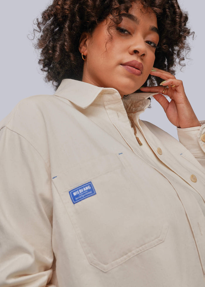 1X=A close-up of a woman wearing a light beige button-up shirt jacket with a blue 'WILDFANG' brand patch on the chest pocket, set against a plain light gray background.