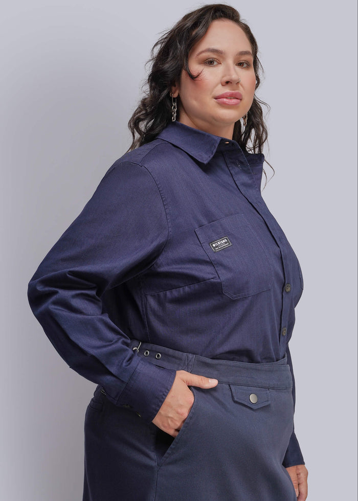 1X=A woman wearing a dark navy blue long-sleeve button-up shirt with a chest pocket and a matching dark skirt, posed against a solid light gray studio background.