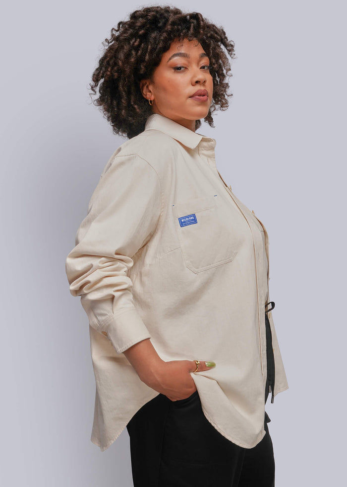 1X=A woman with curly dark hair models a light beige long-sleeve button-up shirt with a chest pocket and a small blue brand tag, paired with black pants against a light gray studio background.