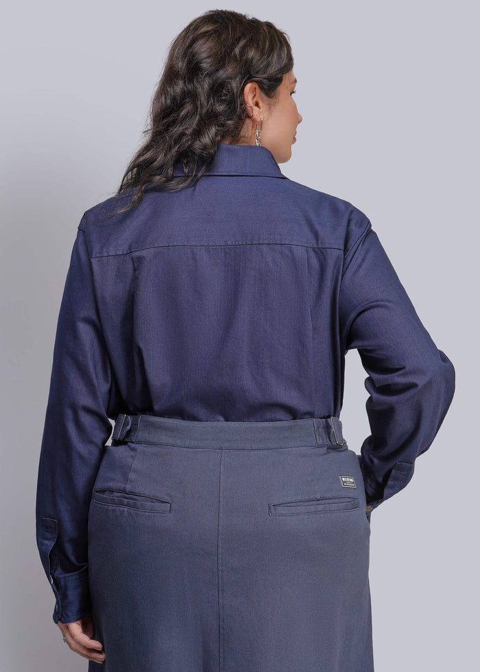 1X=A woman shown from the back wearing a dark navy blue long-sleeve collared shirt tucked into a matching navy skirt, against a solid light gray studio background.