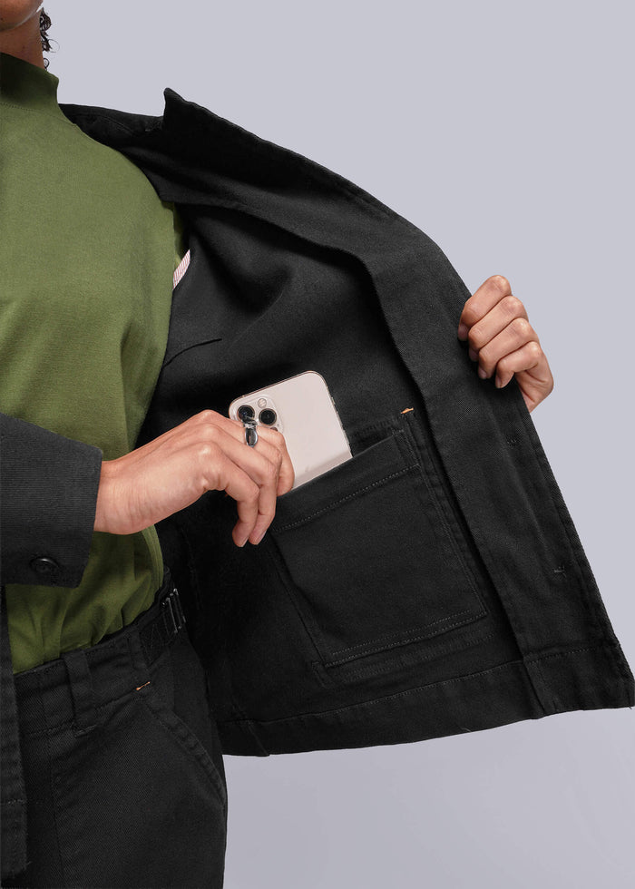 S=Person wearing a black jacket with a white phone in the pocket against a light gray background