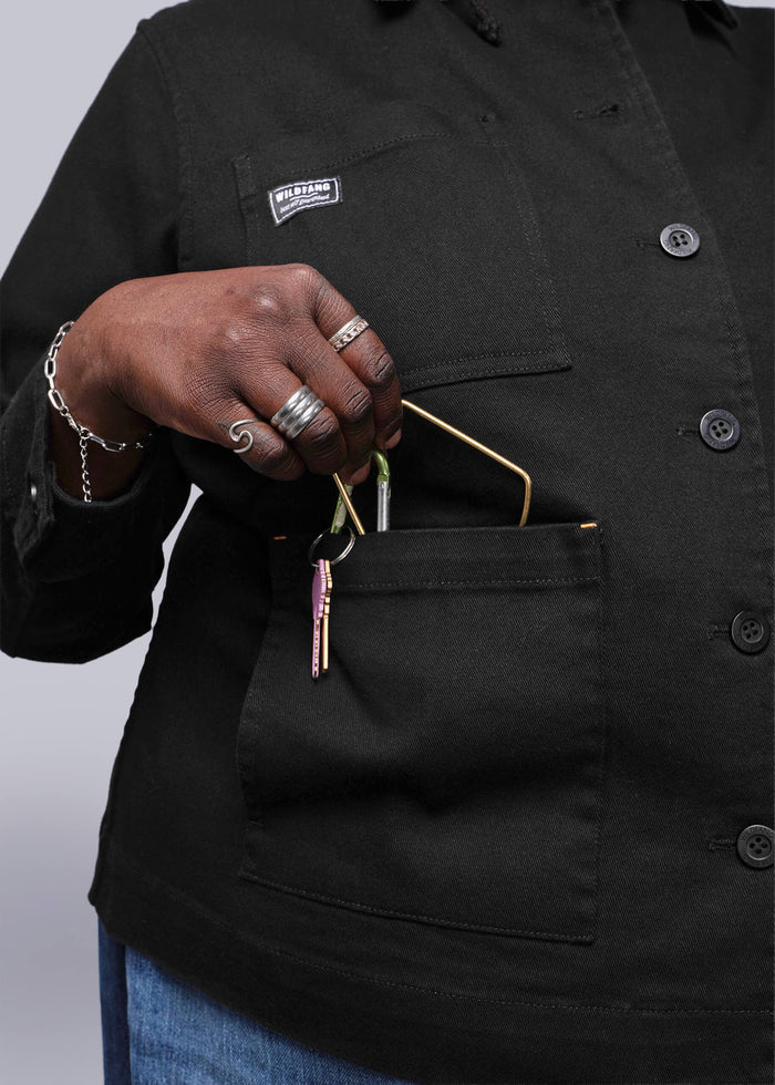 1X=Person wearing a black jacket with a Wildfang logo, placing keys in a pocket, on a light grey background