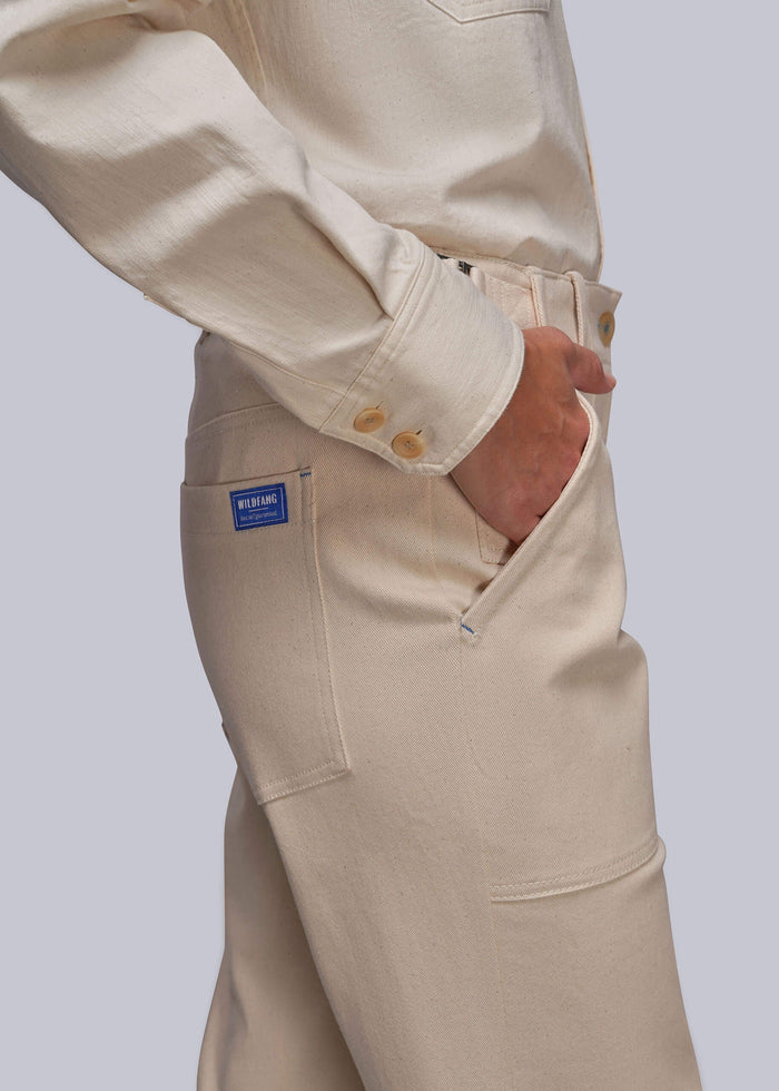 S=Close-up of a person wearing off-white denim work pants with a blue brand patch on the pocket, paired with a matching off-white shirt against a light gray background.