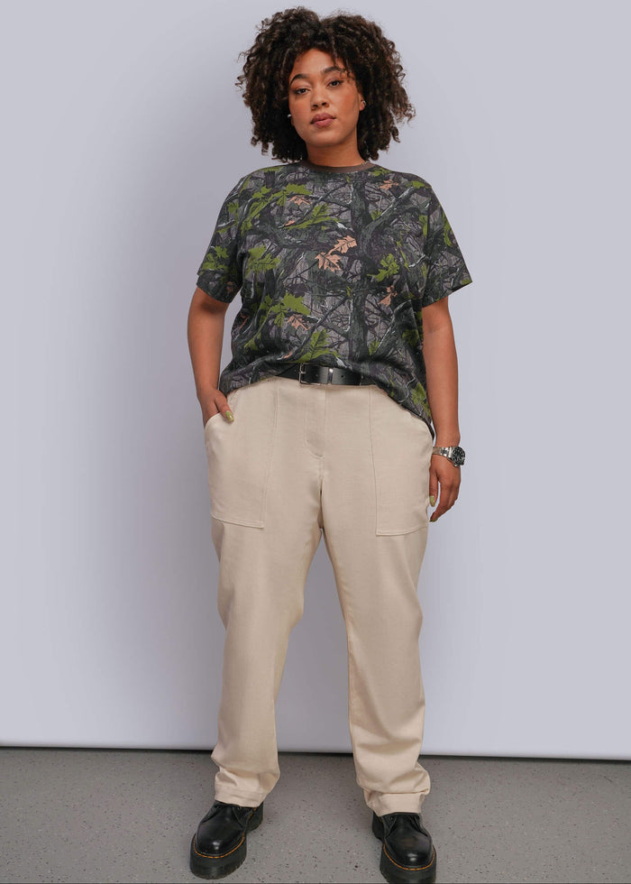 1X=A model stands against a light gray studio wall wearing a short-sleeve crewneck t-shirt with a dark green and black tree camouflage pattern, paired with beige cargo pants and black leather shoes.