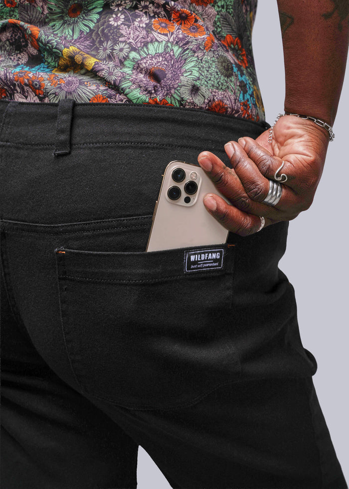 1X=Close up of person wearing black pants with a Wildfang label, putting phone in pocket, against a neutral background