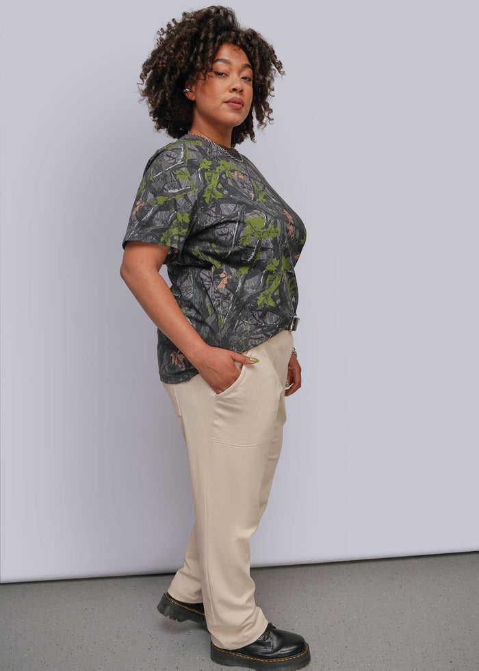 1X=A woman modeling a short-sleeve crewneck t-shirt with a green and gray camouflage pattern, paired with beige trousers and black shoes, standing against a light gray studio background.