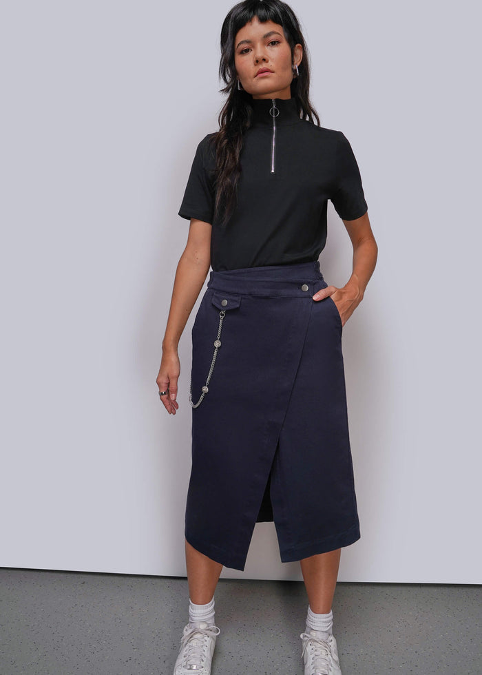 S=A model stands against a light gray studio wall wearing a black short-sleeve mock-neck top with a silver zipper and a dark navy wrap-style midi skirt with silver chain details. She is also wearing white socks and white sneakers.