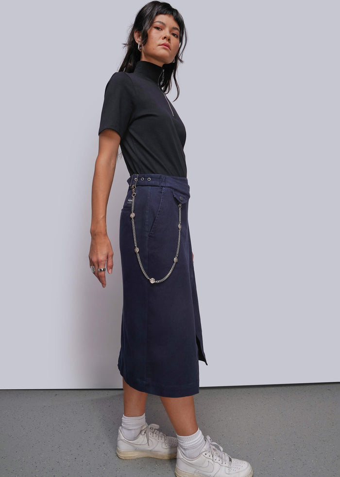 S=A model wearing a black short-sleeve mock neck top and a dark navy midi skirt with a silver chain detail, paired with white socks and sneakers, standing against a light gray studio background.