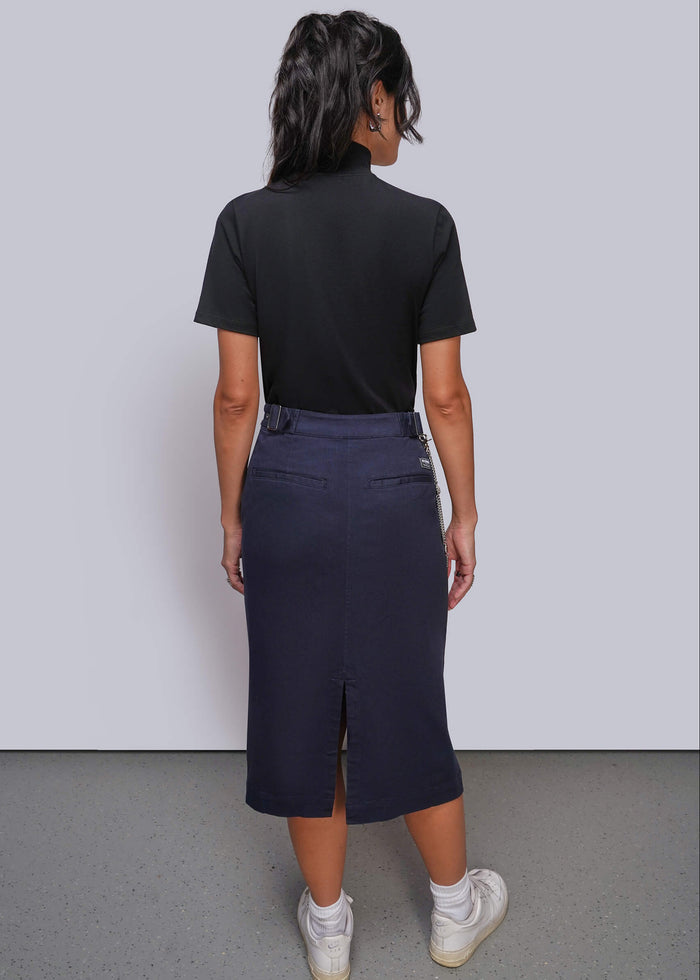 S=A woman is shown from behind wearing a black short-sleeve mock neck top and a dark navy blue midi skirt with a slit, paired with white sneakers, against a light gray studio background.