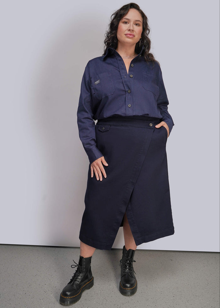 1X=A woman modeling a dark navy blue button up shirt and matching dark navy wrap midi skirt, paired with black combat boots, against a light gray studio background.