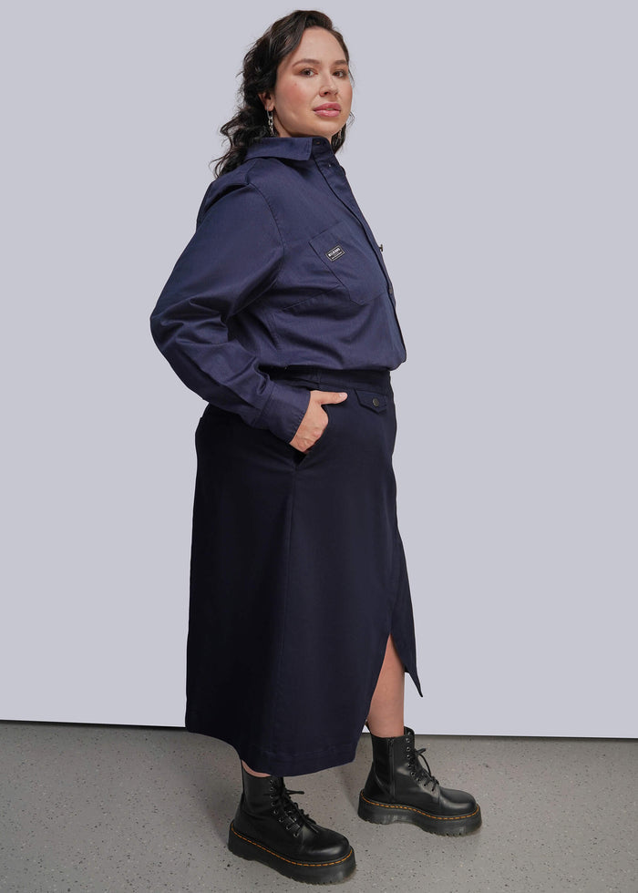 1X=A woman modeling a navy blue button up and matching wrap skirt set, paired with black combat boots, against a light gray studio background.