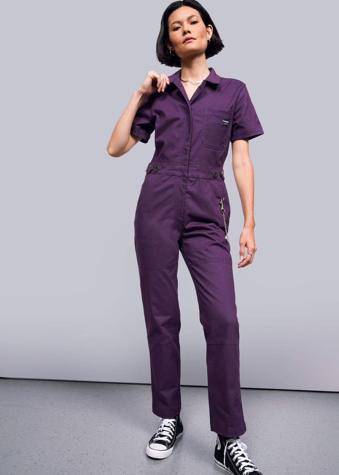 S=A person standing in a studio setting, wearing a short-sleeved purple coverall with a collar, buttons at the waist, and long pants.