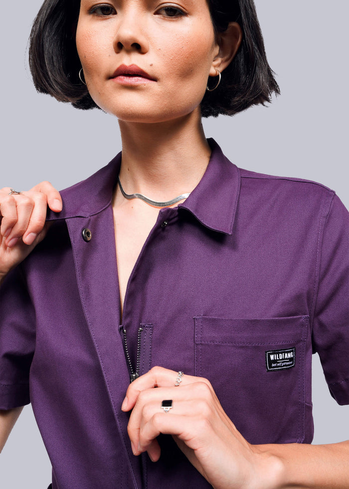 S=Close up of person wearing a purple jumpsuit with a brand logo on a gray background