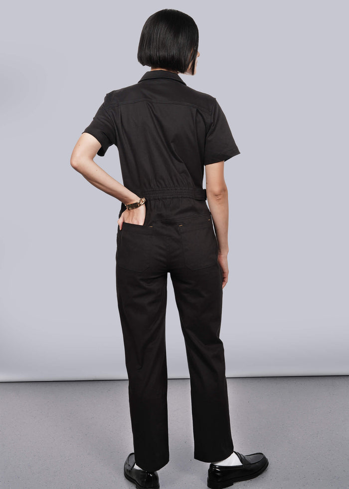 S= A person with short dark hair, wearing The Essential High Waisted Coverall in black—featuring a high-waisted fit and deep pockets—stands against a plain light background, hand on hip, paired with black shoes and white socks.
