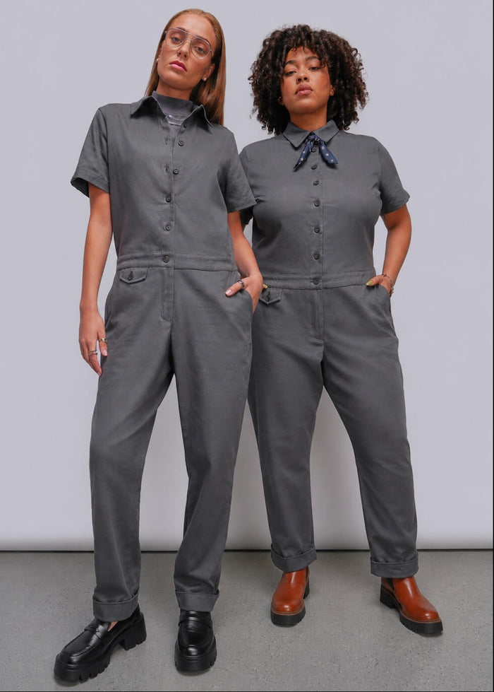Two models wearing matching gray short-sleeve utility jumpsuits, standing against a light gray studio background. The jumpsuit on the left features a button-front chest and a dark gray shirt underneath, paired with black shoes.