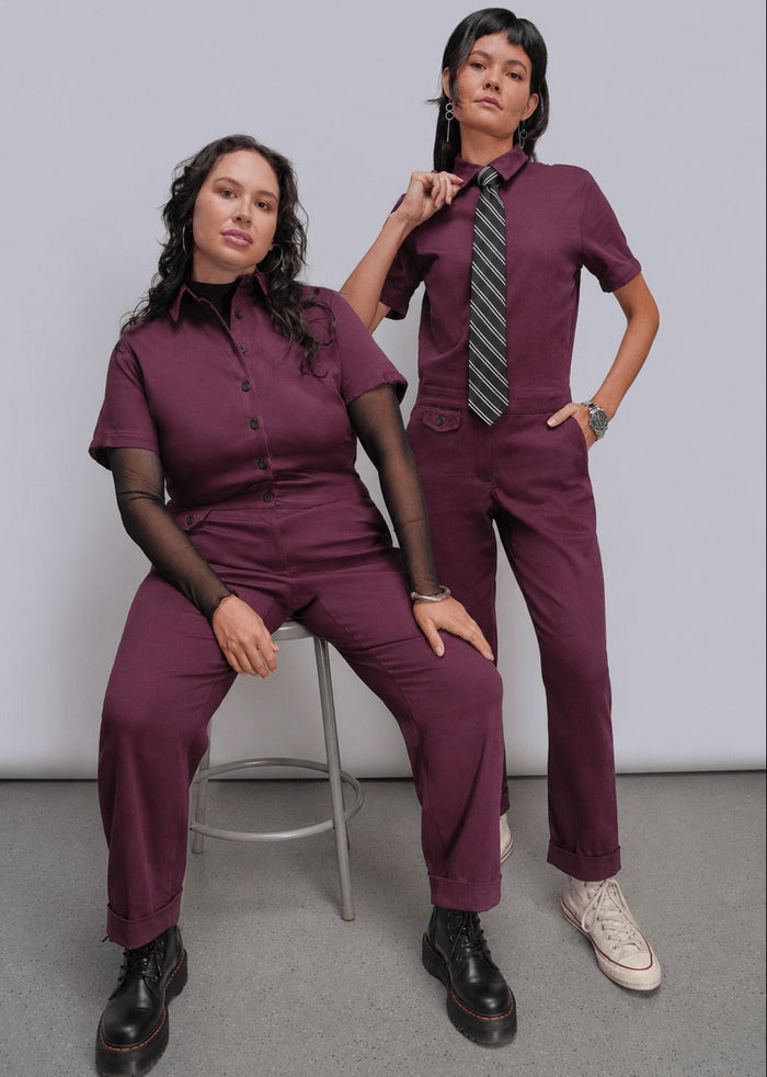 Two women modeling matching burgundy jumpsuits in a studio setting. One woman sits on a metal stool wearing a short-sleeve button-front scrub top and matching pants, while the other stands wearing the same uniform with a striped tie and white sneakers.