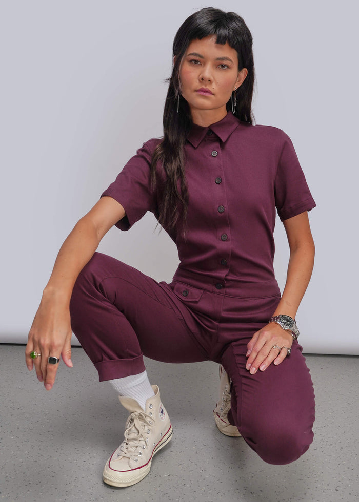 S=A model crouches in a studio wearing a matching short-sleeve button-front shirt and trousers set in a deep plum purple color, paired with white socks and beige high-top sneakers.
