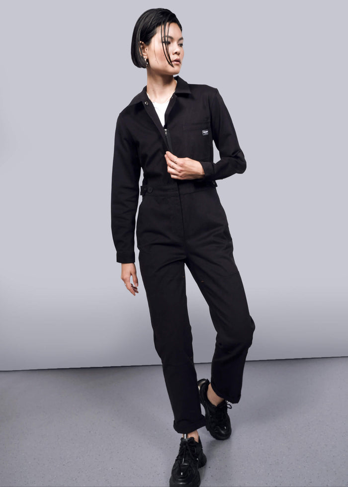 S=A person with short, wet-look hair models The Essential Long Sleeve High Waisted Coverall in black with deep pockets and black sneakers, standing against a plain white background, looking sideways with one hand on the zipper.