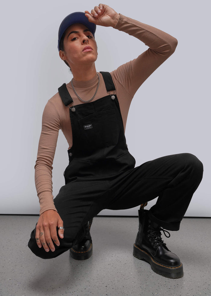 S=A model crouches in a studio wearing black overall-style bib pants over a beige long-sleeve top, with black boots, a dark cap, and silver chain necklaces against a light gray background.