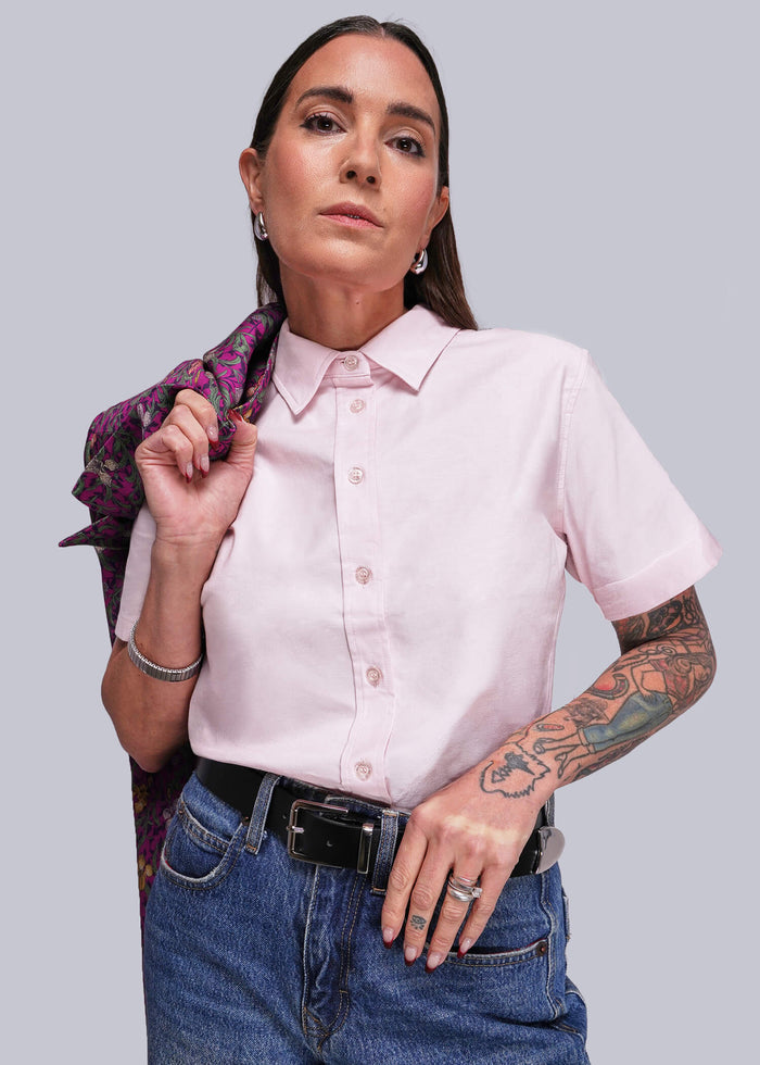 S=Woman wearing a pink shirt and blue jeans with a patterned blazer over her shoulder, standing against a plain background.