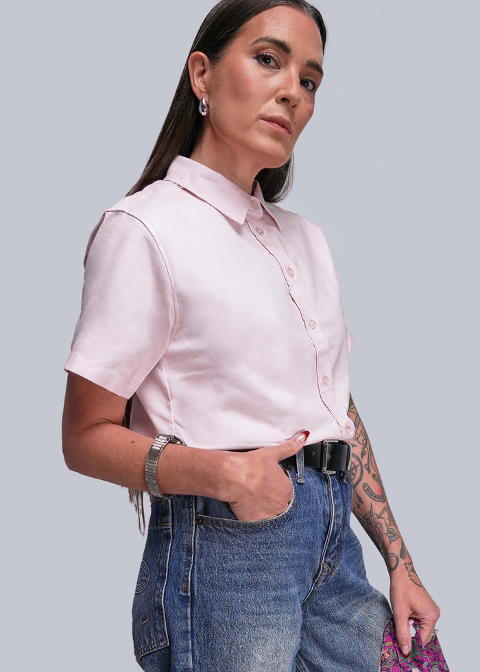 S=Woman wearing a pink shirt, blue jeans, and a black belt standing against a plain background.