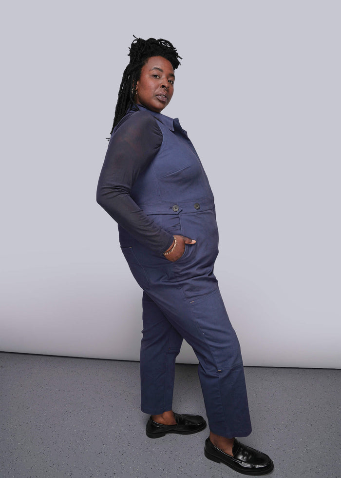 1X=Person wearing a blue coverall with hand in side pocket against a plain background