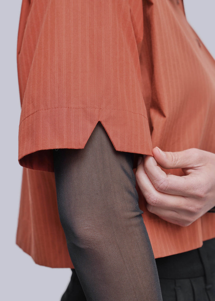 S=Close-up of a person wearing a short-sleeve terracotta orange pinstriped shirt with a hand adjusting the hem, paired with sheer black tights and dark trousers, against a light gray background.