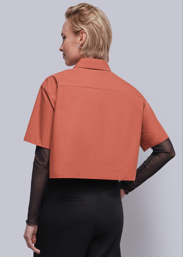 S=A woman shown from the back wearing a short-sleeve cropped collared shirt in a muted coral-orange color with subtle vertical texture, layered over sheer black long sleeves and black trousers, against a solid light gray studio background.