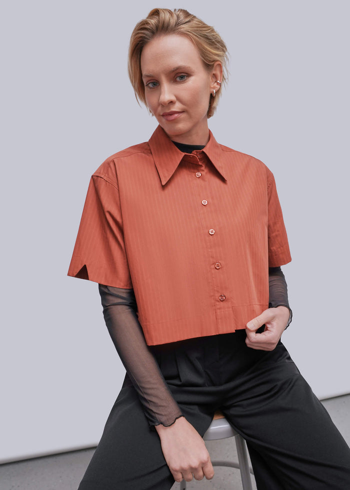 S=A woman seated on a stool wearing a short-sleeve collared button-front top in a muted coral-orange tone over sheer black long-sleeve sleeves, paired with black pants, against a plain light gray studio background.