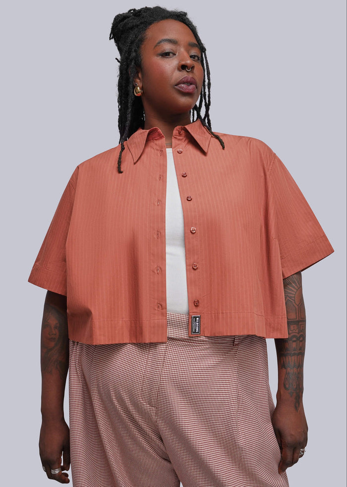 1X=A model wearing a short-sleeve cropped button-up shirt in a muted coral pink, styled over a white t-shirt and paired with herringbone patterned trousers, against a solid gray studio background.