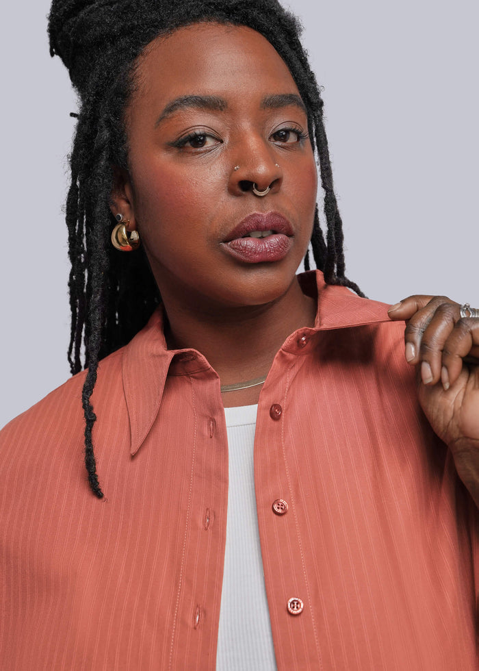 1X=A close-up shot of a model wearing a coral pink button-down shirt with a subtle vertical texture, layered over a white top. The model is shown against a solid light gray background.