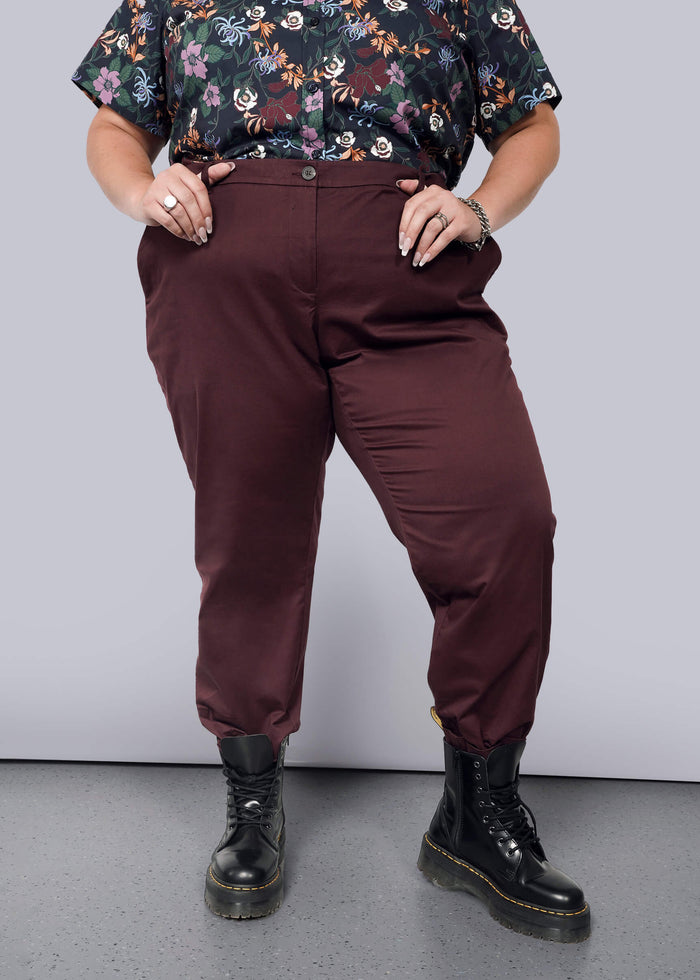 1X=A person in a floral shirt and The Essential Trouser with a burgundy, tapered leg stands with hands in pockets. They wear black boots on a grey speckled floor against a plain white background.