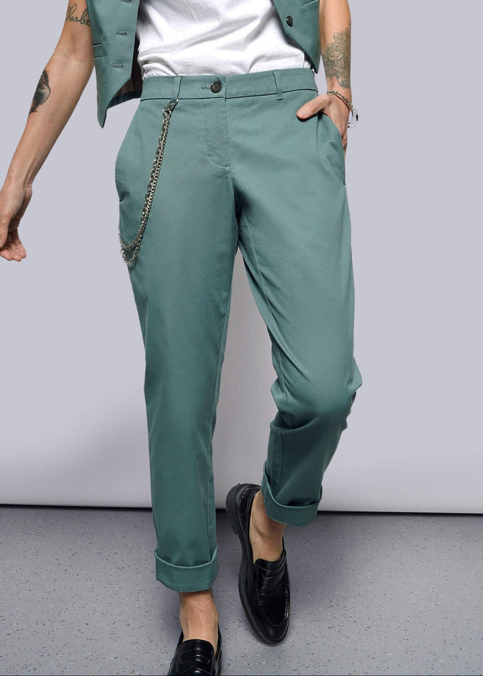 S= A person stands casually with a tattooed arm, hand in their pocket, wearing The Essential Trouser in classic teal with a chain accessory, paired with a white shirt and cropped teal vest. They wear black loafers against a plain white background.