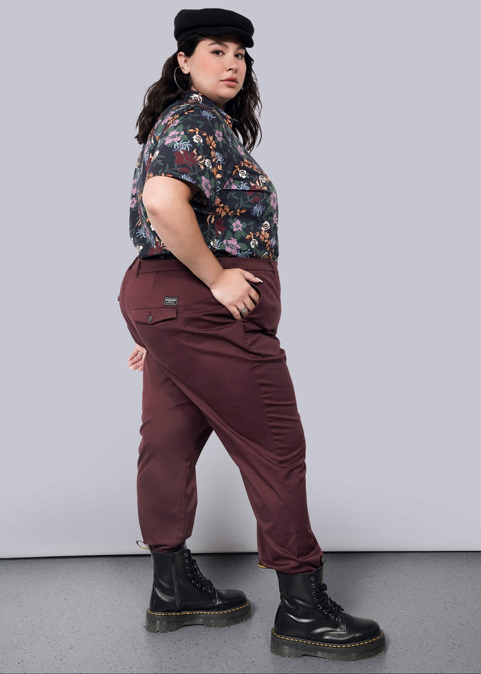 1X=A person stands confidently against a white background, hands in pockets, slightly turned to the side. They wear a black cap, floral shirt, The Essential Trouser in maroon with a tapered leg, and black boots.