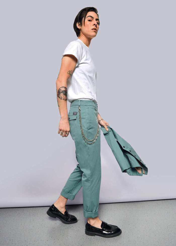S=A tattooed person in a white t-shirt, The Essential Trouser in teal with a chain, black shoes, and holding a matching teal blazer walks against a plain white background.