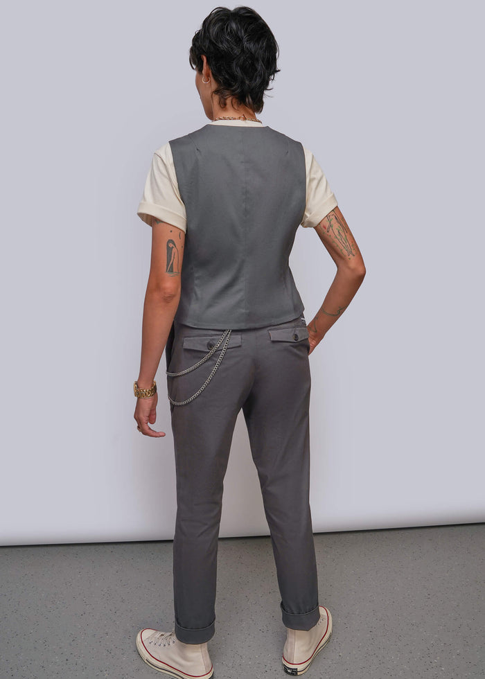 S=A model shown from the back wearing a dark gray sleeveless vest over a cream t-shirt, dark gray trousers with a chain detail at the back pocket, and beige sneakers, set against a plain light gray studio background.