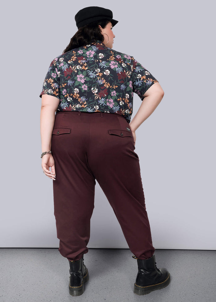 1X=Standing against a plain white background, a person faces away wearing The Essential Trouser in classic maroon with a tapered leg, paired with a floral shirt and black cap. Black boots complete the ensemble as they pose with one hand on their hip.