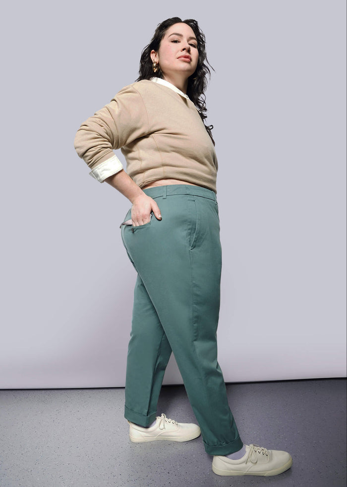 1X=A woman poses confidently in a studio against a white background, wearing The Essential Trouser in teal green with a relaxed mid-rise fit, paired with a beige sweater and white sneakers. She has dark wavy hair and one hand in her pocket.